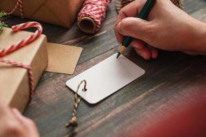 Frau, die Geschenkanhänger schreibt und an Weihnachtsgeschenk auf Holztisch anbringt foto