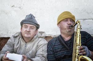 Obdachlose Straßenmusikanten sitzen mit einem Saxophon und einer Spendenbox auf der Straße. foto