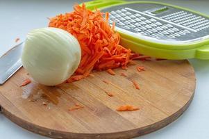 geschälte Zwiebeln, geriebene Karotten und eine Reibe auf einem Schneidebrett. Zutaten für Suppe auf weißem Hintergrund. foto