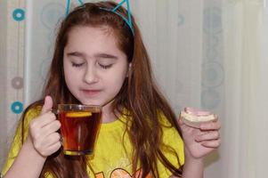 Ein junges Mädchen sitzt an einem Tisch in der Küche und trinkt Tee mit einem Sandwich. Morgen, Frühstück, Junkfood. foto