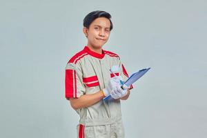 Fröhlicher junger männlicher Mechaniker, der Notizen über die Zwischenablage auf grauem Hintergrund macht foto