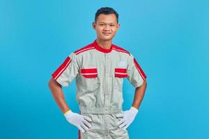 Porträt eines lächelnden jungen asiatischen Mechanikers, der selbstbewusst die Hände über blauem Hintergrund hält foto