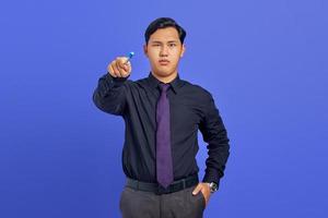 Ernsthafter, gutaussehender junger Geschäftsmann, der einen Stift hält und auf die Kamera auf violettem Hintergrund zeigt foto