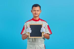 Porträt des fröhlichen jungen asiatischen Mechanikers, der leeres Brett auf blauem Hintergrund zeigt foto