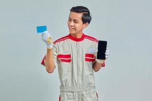 Porträt eines lächelnden jungen Mechanikers, der die Kreditkarte in der Hand betrachtet und das Smartphone auf grauem Hintergrund hält foto
