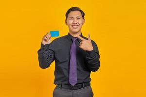Porträt eines lächelnden jungen asiatischen Geschäftsmannes, der mit dem Finger auf die Kreditkarte auf gelbem Hintergrund zeigt foto