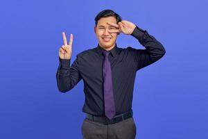 Foto eines lächelnden gutaussehenden Geschäftsmannes, der ein Friedenszeichen über den Augen auf violettem Hintergrund zeigt