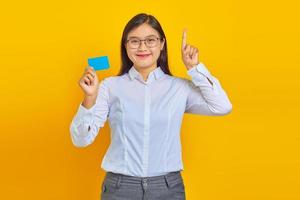 Foto lächelnde asiatische Frau, die Kreditkarte hält und auf gelbem Hintergrund nach oben zeigt