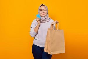 Porträt einer lächelnden asiatischen Frau, die Einkaufstasche hält und Kreditkarte auf gelbem Hintergrund zeigt foto