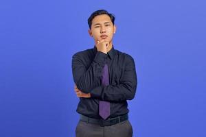 Nachdenklicher junger gutaussehender Geschäftsmann reibt sich das Kinn und denkt über eine Frage auf violettem Hintergrund nach foto