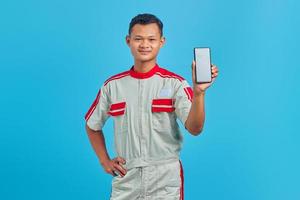 Porträt eines lächelnden jungen Mechanikers, der einen leeren Smartphone-Bildschirm in der Hand zeigt, isoliert auf blauem Hintergrund foto