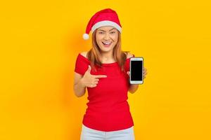 fröhliche junge asiatische frau in weihnachtsmannmütze, die mit dem finger auf ein leeres handy auf gelbem hintergrund zeigt