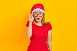 überraschte junge asiatische frau mit weihnachtsmütze mit brille bei offenem mund auf gelbem hintergrund foto