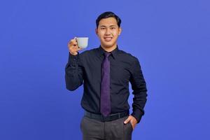 hübscher junger Geschäftsmann, der mit den Händen in der Tasche in die Kamera lächelt und einen Becher auf violettem Hintergrund hält foto