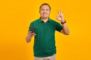 lächelnder junger Mann, der Smartphone benutzt und eine gute Geste auf gelbem Hintergrund macht foto