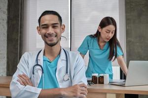 Porträt des jungen männlichen Arztes asiatischer Abstammung in Uniform mit Stethoskop. Lächeln und Blick in die Kamera in einer Krankenhausklinik, weibliche Partnerin, die dahinter arbeitet, Zwei-Personen-Medikamentenfachmann. foto