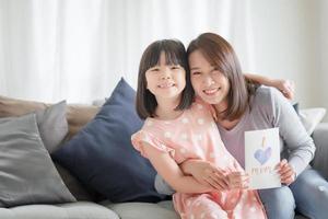 Asiatische Mutter umarmt ihre süße Tochter, die eine handgemachte Grußkarte mit dem Wort "I Love Mom" gibt, um sie zu Hause zu überraschen foto