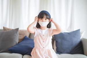 Asiatisches kleines süßes Mädchen, das eine hygienische Gesichtsmaske trägt, um den Ausbruch von Coronavirus oder Covid-19 zu verhindern, halten soziale Distanz und bleiben zu Hause.