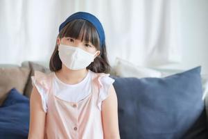 Asiatisches kleines süßes Mädchen, das eine hygienische Gesichtsmaske trägt, um den Ausbruch von Coronavirus oder Covid-19 zu verhindern, halten soziale Distanz und bleiben zu Hause.