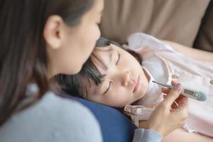 Asiatische Mutter, die die Körpertemperatur ihres Kindes mit einem Thermometer überprüft, das zu Hause Fieber und Krankheit bekam.