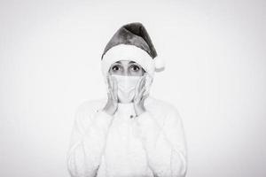 Frau mit rotem Weihnachtshut auf überraschten Händen, um die mit Gesichtsmaske bedeckten Wangen zu hetzen. Konzept der Nachrichtenreaktion und Erstaunen. foto