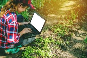 junge asiatische Frau mit Laptop im Gemüsegarten der Parzellen sitzen. Verwendung von Agrartechnologie Verwendung von Tablet-Computer-Analysedaten Lesen eines Berichts.