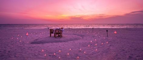 Schöner Tisch für ein romantisches Essen am Strand mit Laternen und Stühlen und Blumen mit Kerzen und Himmel und Meer im Hintergrund. Abendessen am Strand bei Sonnenuntergang