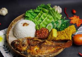 eine Packung Reis, gebratener Fisch und etwas frisches Gemüse auf schwarzem Hintergrund