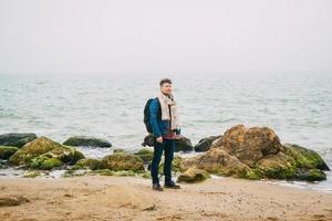 Reisender Mann mit Rucksack, der am Sandstrand inmitten von Felsen vor Meereshintergrund steht foto