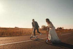 lächelndes Hochzeitspaar, das bei Sonnenuntergang auf Rollern entlang der Straße außerhalb der Stadt fährt. Platz für Text oder Werbung foto