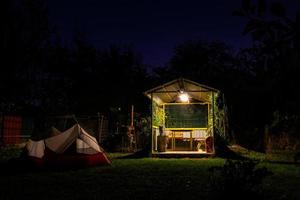 Zelte gegen Unterschlupf. Campingplatz mit weiß-rotem Zelt und Mini-Shelterhaus auf dem Campingplatz, der nachts von Natur umgeben ist. keine Person. foto