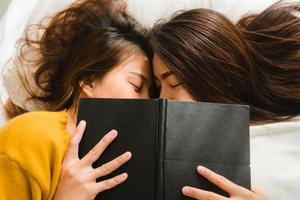 Draufsicht auf schöne junge asiatische Frauen lesbisches glückliches Paar küssen und lächeln, während sie zu Hause zusammen im Bett unter Buch liegen. lustige Frauen nach dem Aufwachen. Lesbenpaar zusammen drinnen Konzept.