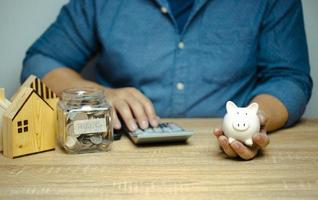 Mann mit Sparschwein, der den Taschenrechner drückt, berechnet Einnahmen und Ausgaben auf braunem Holztisch, Münzsparschwein, Anlageideen, Sparen, um ein Haus zu kaufen foto