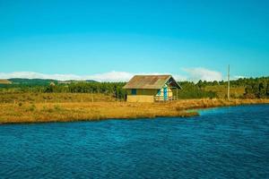 cambara do sul, brasilien - 18. juli 2019. Holzchalet mit Blick auf einen blauen See in einer hügeligen Landschaft mit Bäumen und trockenen Büschen in der Nähe von cambara do sul. foto