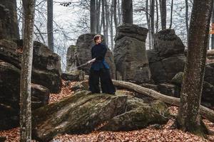 Mann im schwarzen Kimono, der Kampfkunst mit einem Schwert auf Felsen und Waldhintergrund praktiziert foto