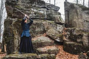 Mann im schwarzen Kimono, der Kampfkunst mit einem Schwert auf Felsen und Waldhintergrund praktiziert foto