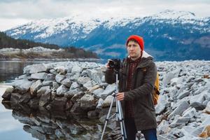 Mann mit Kamera auf Stativ, der auf dem Hintergrund eines Berges und eines Sees steht, macht eine Fotolandschaft foto