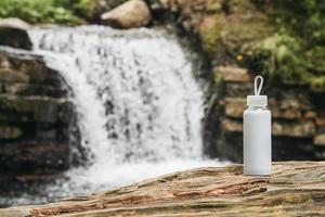 weiße Flasche mit Wasser, die auf einem Holzstamm vor dem Hintergrund eines Flusses und eines Wasserfalls steht foto