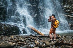 Reisender Mann mit gelbem Rucksack, der auf dem Hintergrund eines Wasserfalls steht, macht eine Fotolandschaft foto