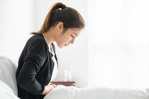 eine frau, die mit unterleibsschmerzen im bett sitzt und ihre hand auf ihren bauch drückt. foto