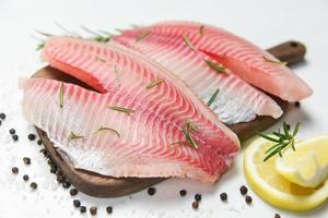 frisches Fischfilet geschnitten für Steak oder Salat mit Kräutergewürzen Rosmarin und Zitrone - roher Tilapiafiletfisch und Salz auf weißem Steinhintergrund und Zutaten zum Kochen von Speisen foto
