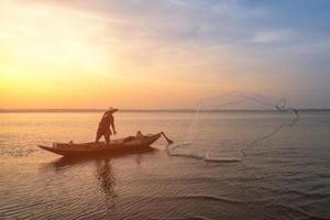 Asiatischer Fischer auf einem Holzboot, das früh während des Sonnenaufgangs ein Netz zum Fangen von Süßwasserfischen im Naturfluss wirft foto