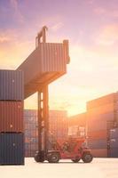 Gabelstapler, der Frachtcontainer im Versandhof oder Dockyard gegen Sonnenaufganghimmel anhebt, mit Frachtcontainerstapel im Hintergrund für Transportimport, -export und logistisches Industriekonzept foto