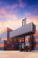 Gabelstapler, der Frachtcontainer im Versandhof oder Dockyard gegen Sonnenaufganghimmel anhebt, mit Frachtcontainerstapel im Hintergrund für Transportimport, -export und logistisches Industriekonzept foto