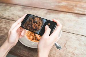 Bloggerin fotografieren Mittagessen im Restaurant mit ihrem Telefon. Eine junge Frau, die Spaghetti-Essen auf dem Smartphone fotografiert und eine Mahlzeit mit einer mobilen Kamera fotografiert. foto