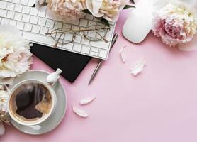 Flacher Damenschreibtisch mit Draufsicht mit Blumen. weiblicher arbeitsplatz mit laptop, blumenpfingstrosen, zubehör, notizbuch, brille, tasse kaffee auf rosa hintergrund.