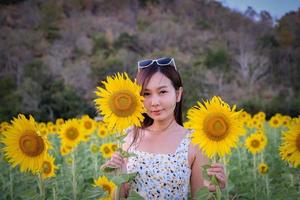 Fröhliches, fröhliches asiatisches Mädchen mit Sonnenblume, das die Natur genießt und im Sommer im Sonnenblumenfeld lächelt. foto