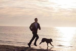 junger Mann, der mit schwarzem Hund am Morgenstrand spazieren geht foto