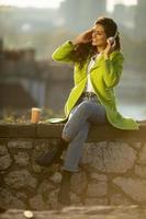 Hübsche junge Frau, die an einem sonnigen Herbsttag Musik mit Smartphone und Kaffee zum Mitnehmen am Fluss hört foto