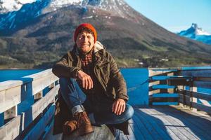 Reisender Mann sitzt auf einem Holzsteg auf einem Bergsee-Hintergrund. Platz für Text oder Werbung